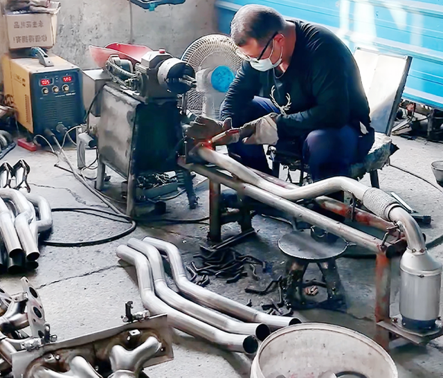 Craftsman welding a car exhaust system, focusing on the catalytic converter and muffler, surrounded by various exhaust pipes and components.